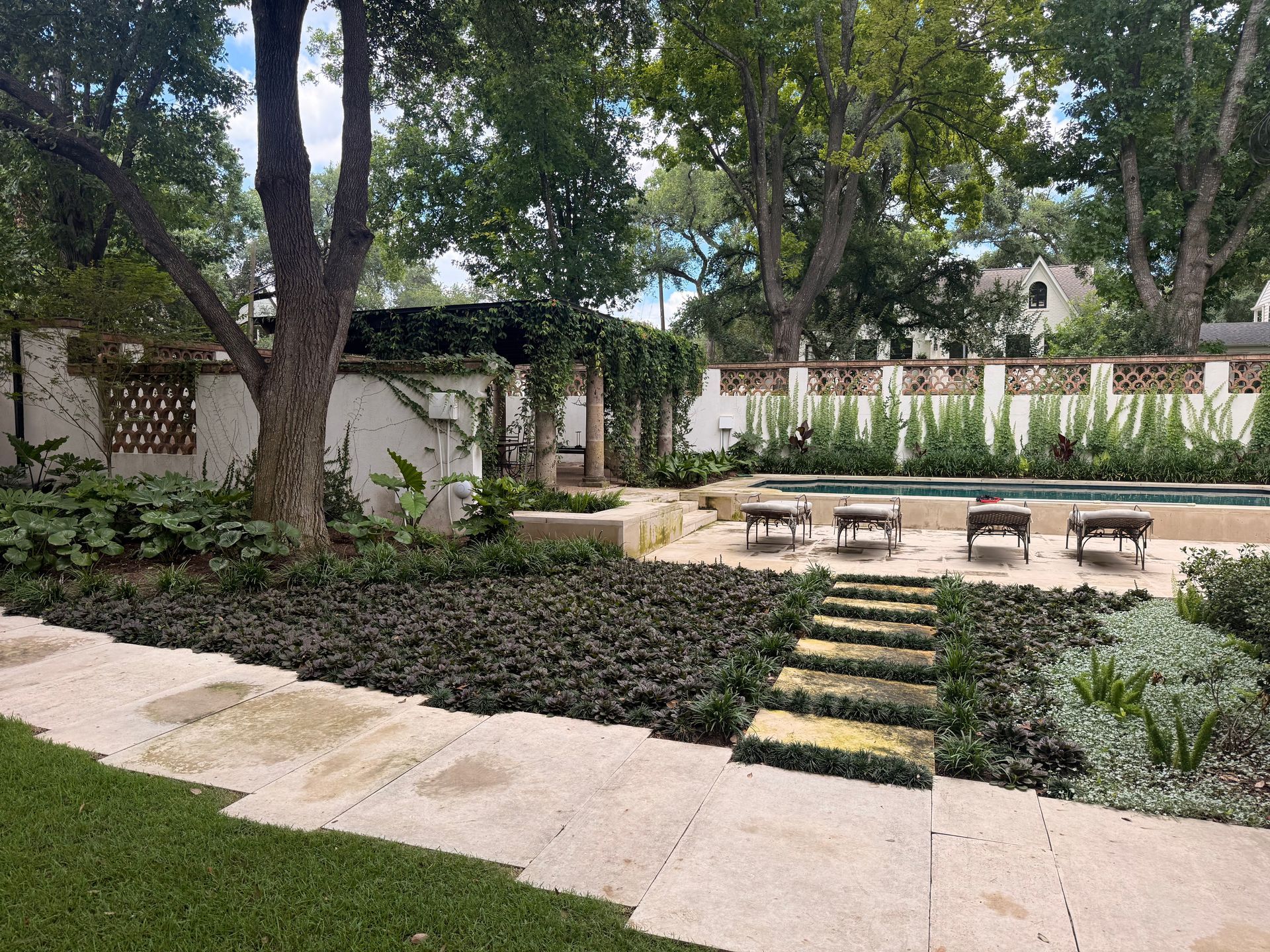 Quiet backyard garden with trees, stone path, dark mulch beds, and seating around a small patio.