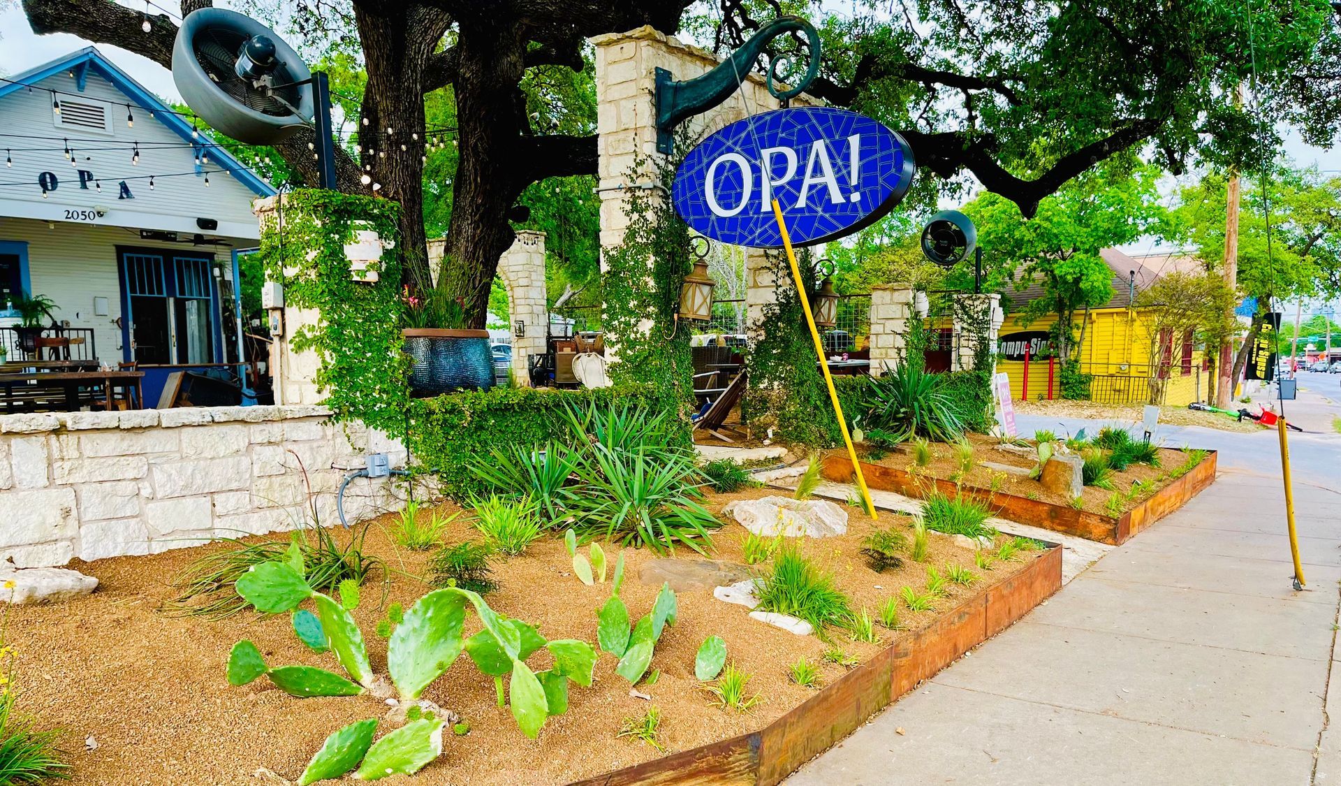 Blue “OPAL” sign by a roadside garden with trees and a white building in the background