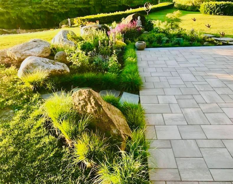 Sunlit garden path beside rocks, green grass, and flowering shrubs