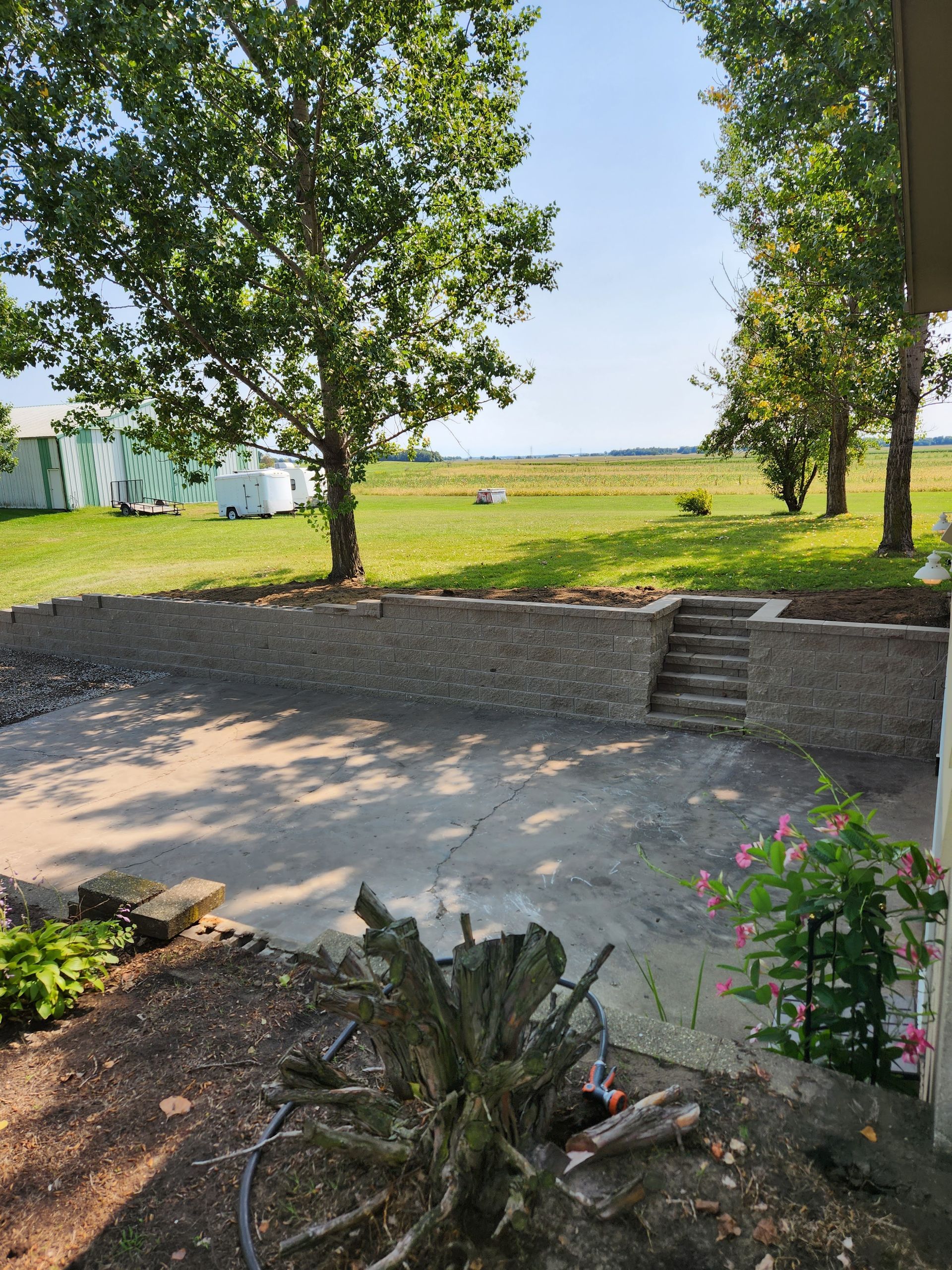 Concrete patio with stone retaining wall, steps leading to a grassy field. Trees frame the view.