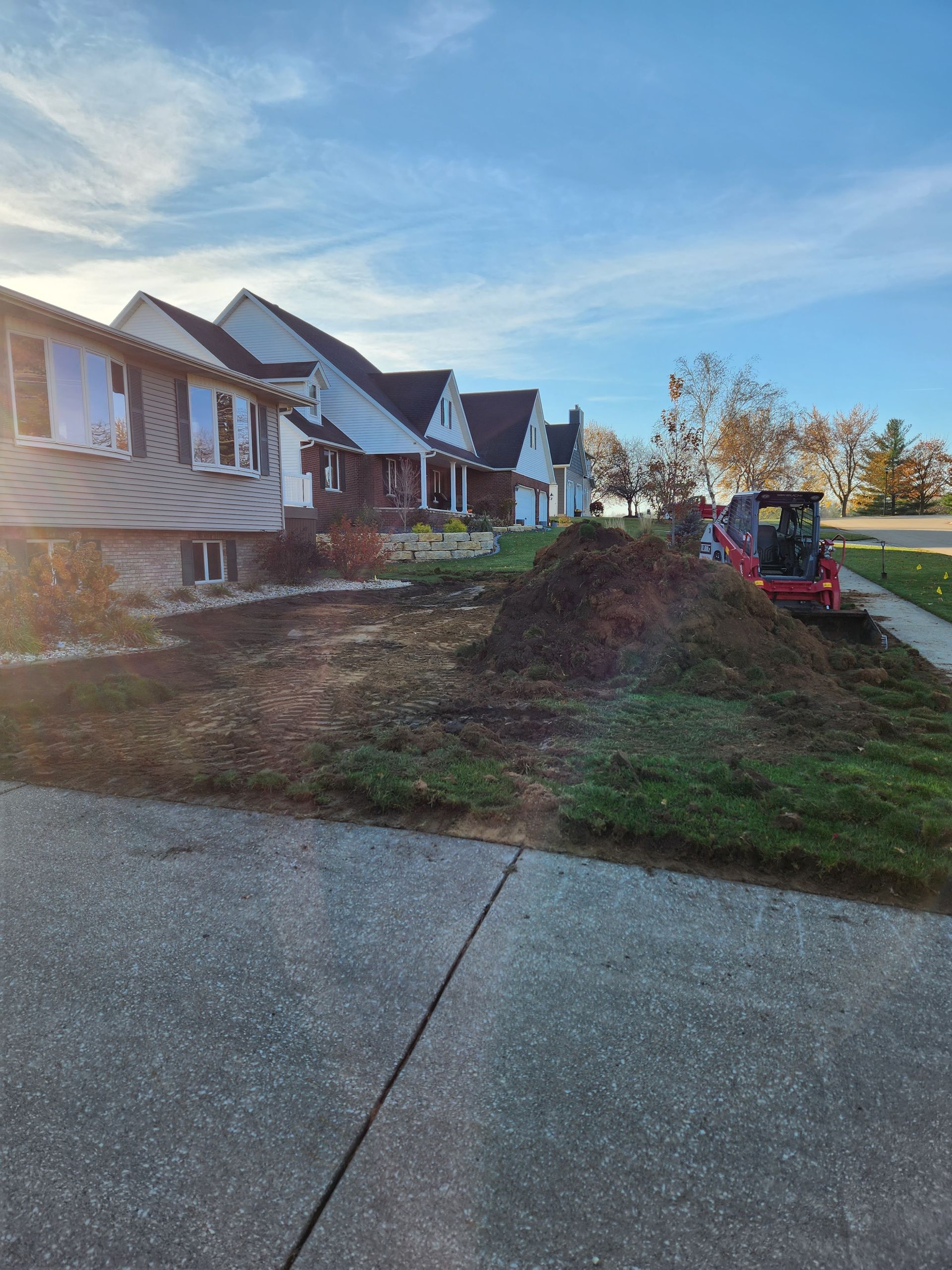 Houses with landscaping under construction; pile of dirt and small excavator on a sunny day.