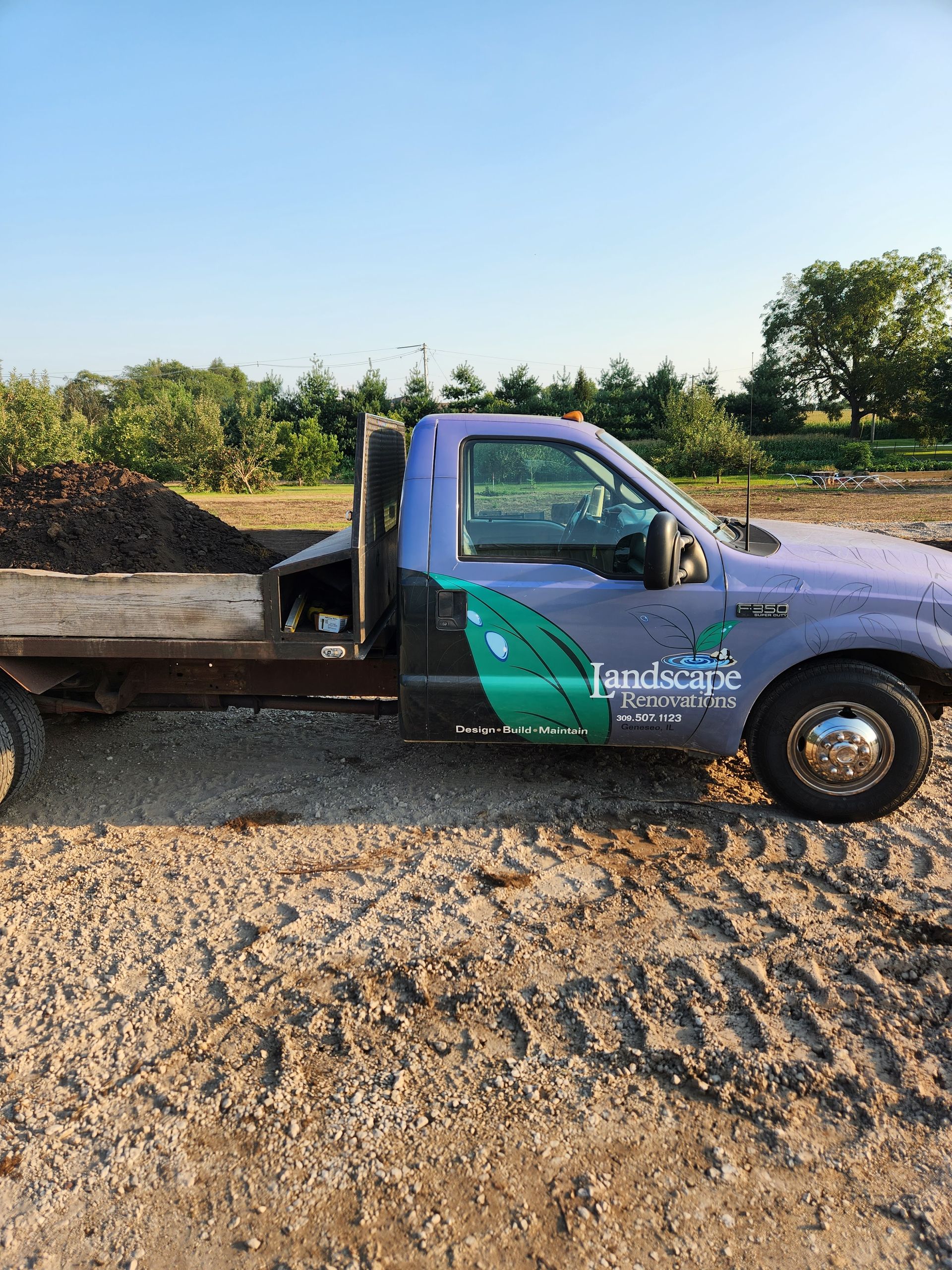 Purple landscape truck filled with dark mulch, parked on dirt.