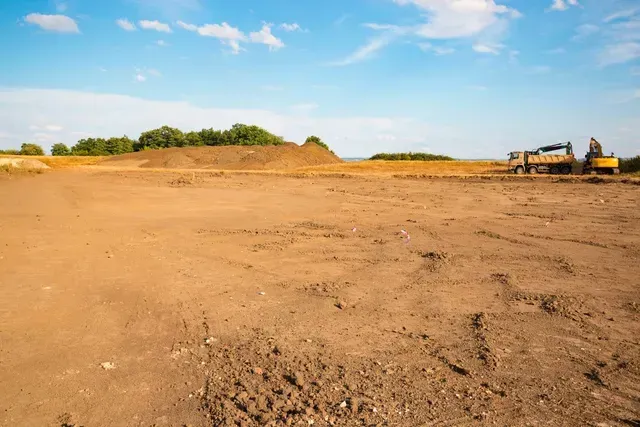 Cleared construction site with dirt, trees in the background, and construction vehicles. Blue sky.