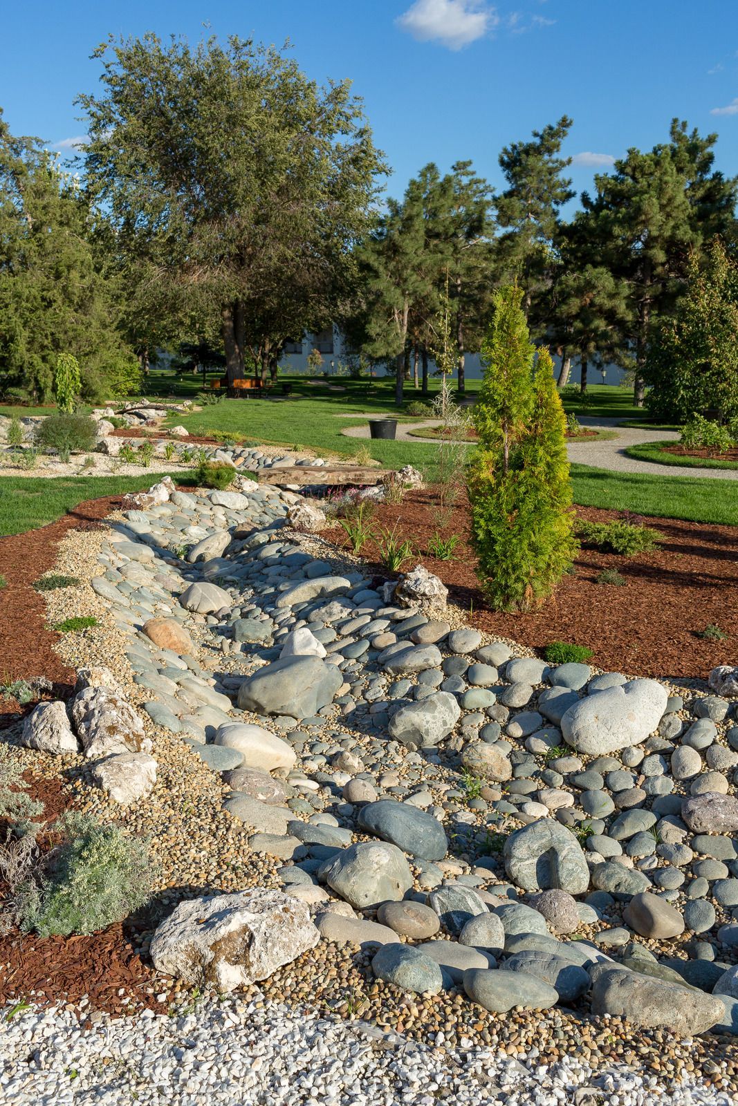 Rocky stream bed in a park, lined with rounded stones and landscaping, under a blue sky.