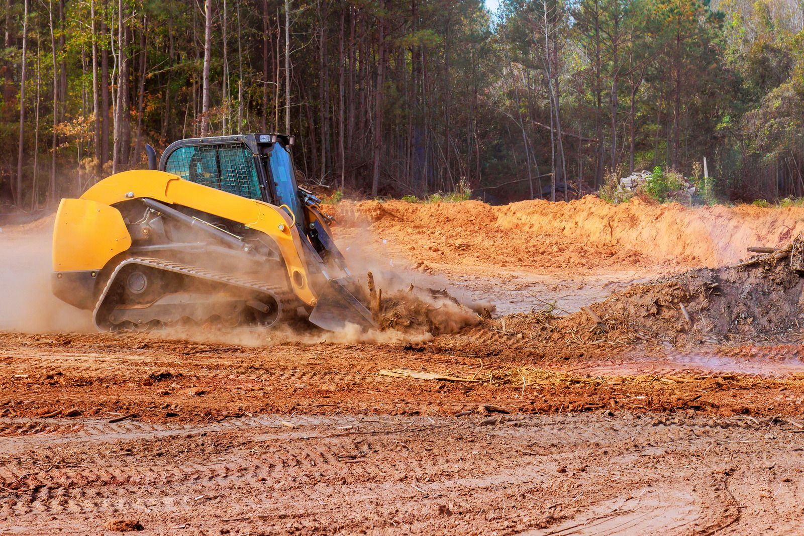 Yellow and blue skid-steer loader moving dirt on a construction site; forest background.