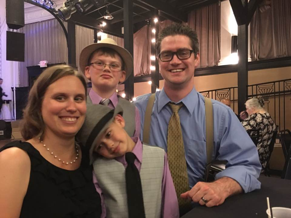 Family of four smiling, posing at an event. Two boys in hats, suspenders, and ties; woman in black dress; man in blue shirt, glasses, and tie.