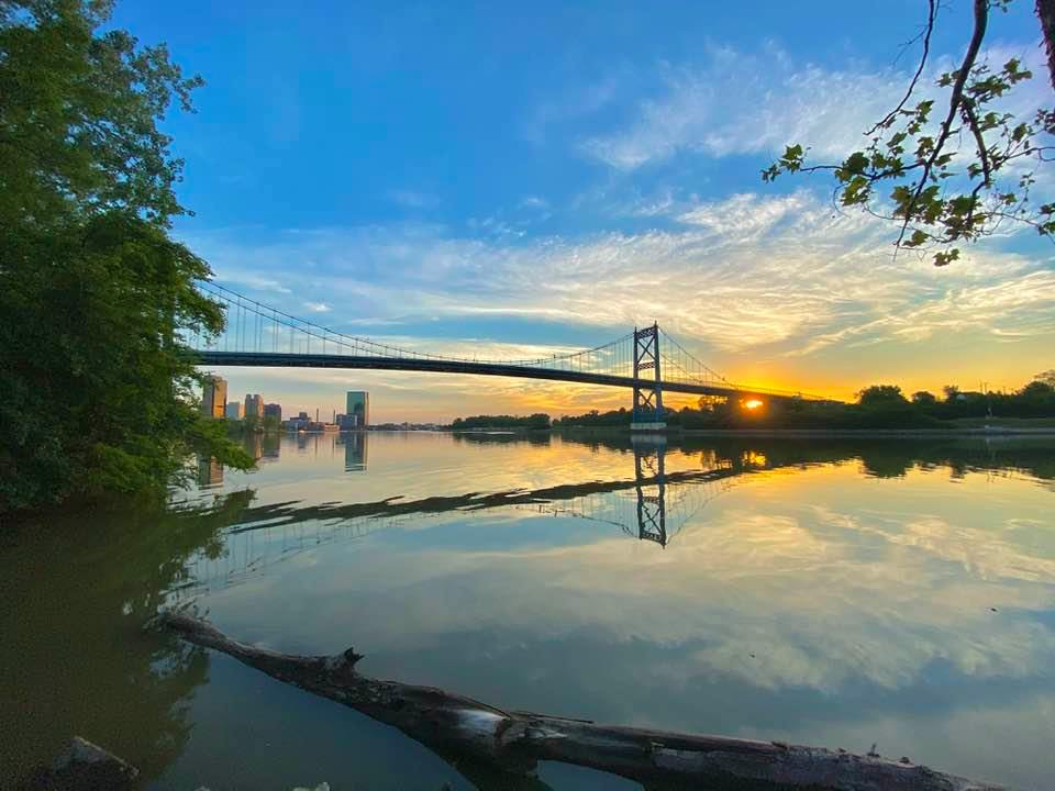 Suspension bridge reflecting in calm water at sunset; city skyline in distance.