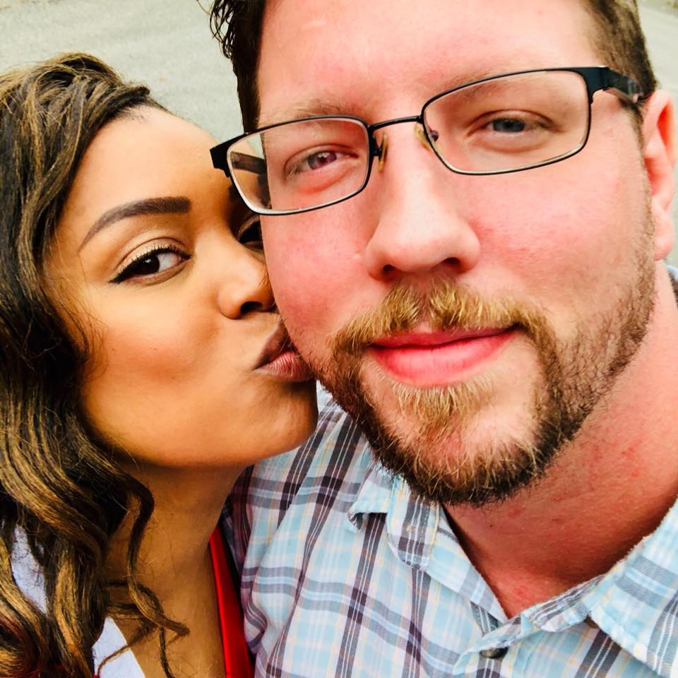 Woman kisses a man's cheek; they are outside. Man wears glasses, beard, plaid shirt.