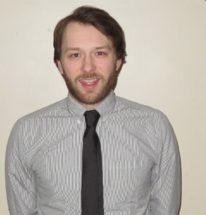 Man in striped shirt and black tie smiles, head and shoulders shot against a beige wall.