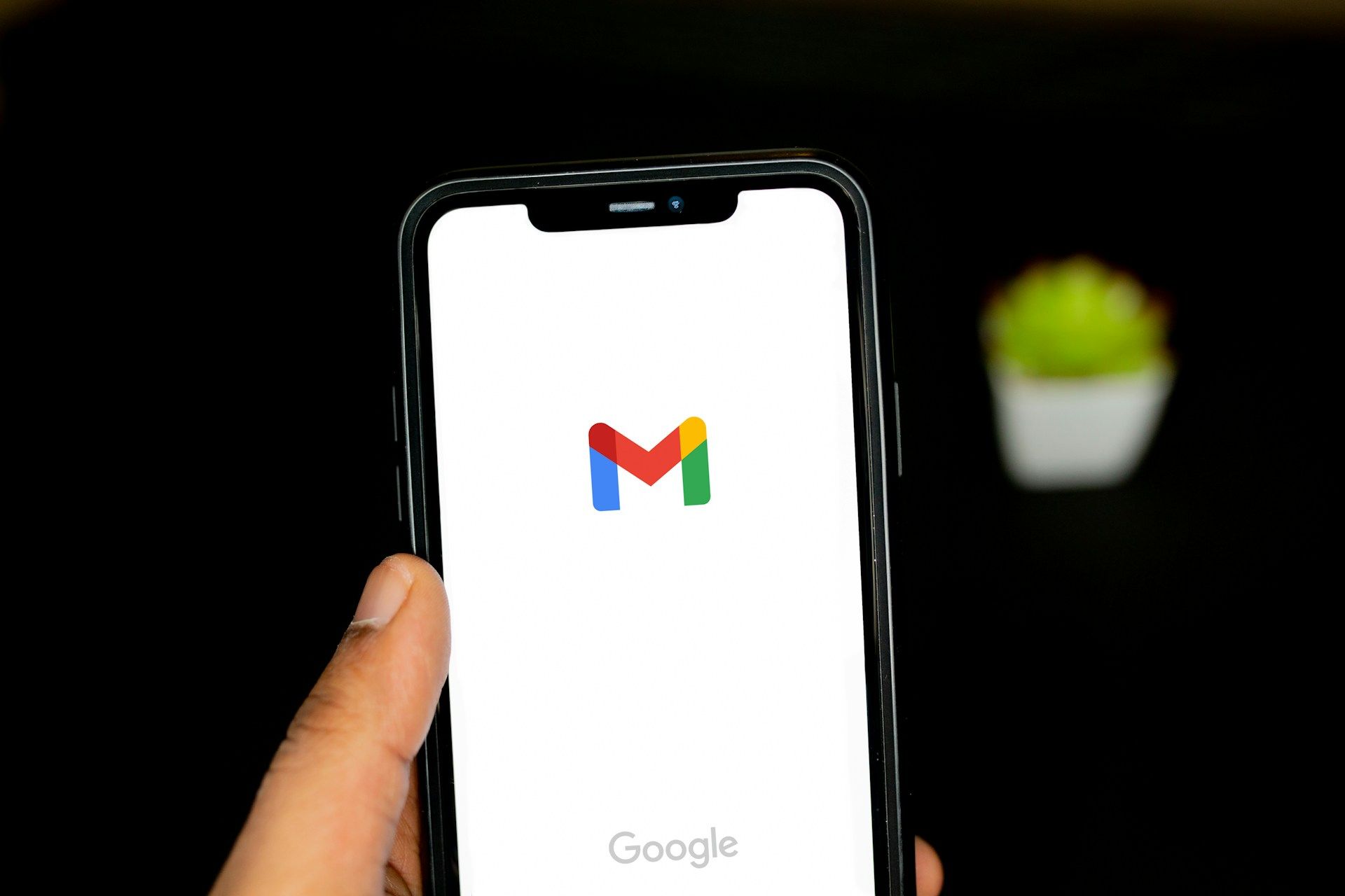 Hand holding smartphone displaying the Gmail logo on a white screen against a dark background.