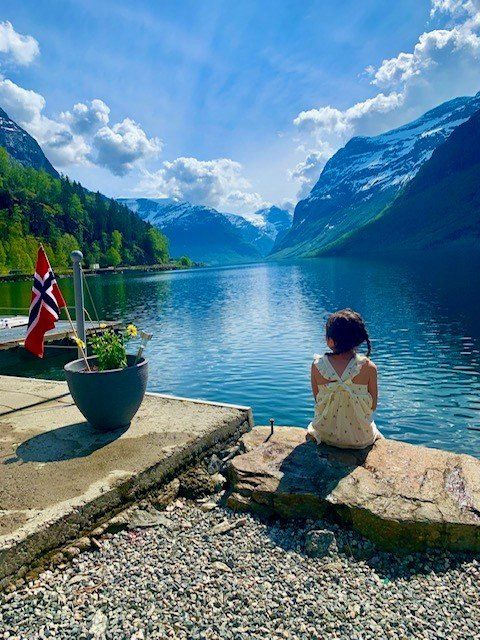 Person som sitter på en steinete innsjø med utsikt over en rolig fjord, med fjell, skyer og et norsk flagg i nærheten.