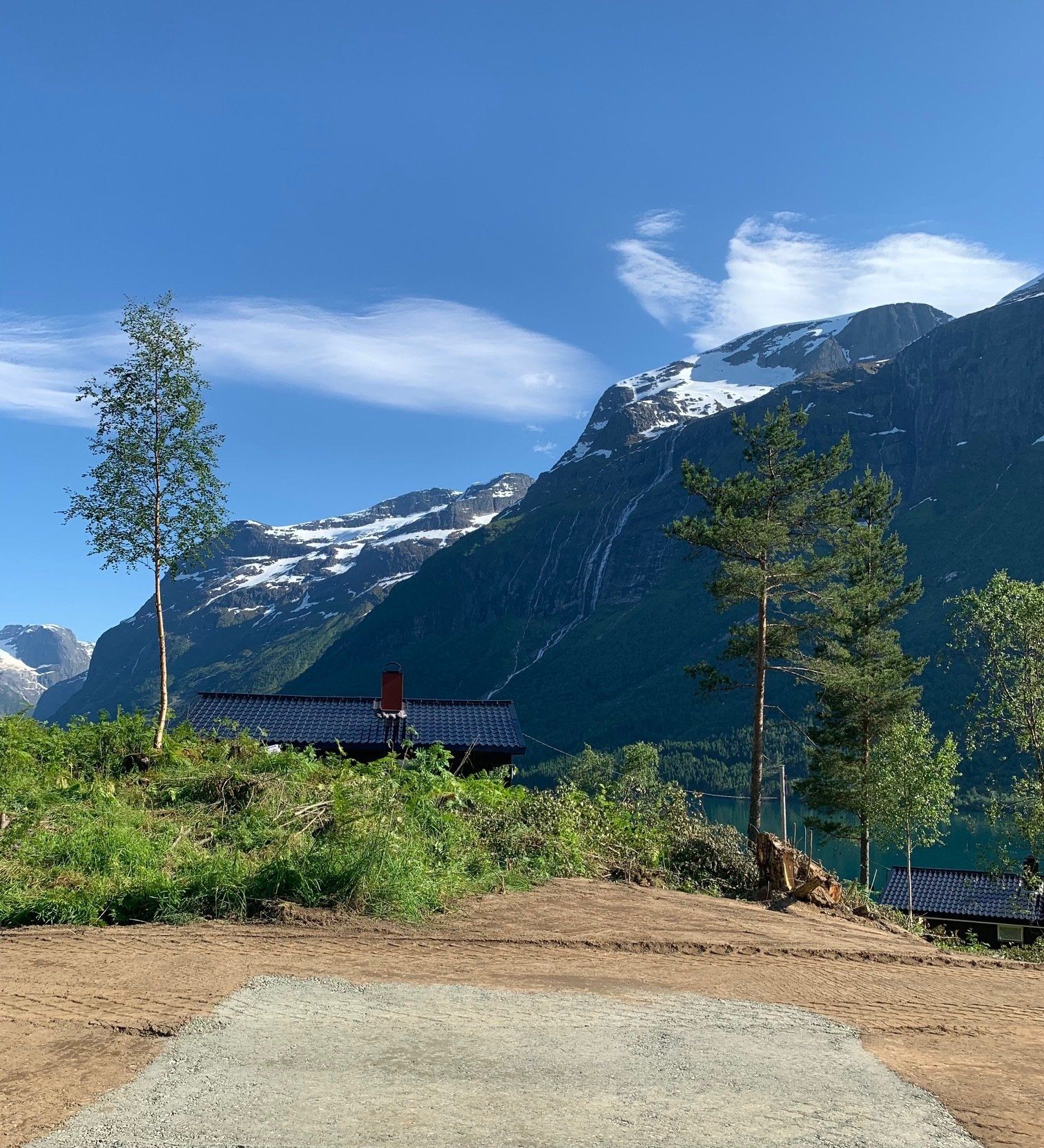 Utsiktspunkt i en fjelldal med blå himmel, snødekte topper, furutrær og en person ved et rekkverk.