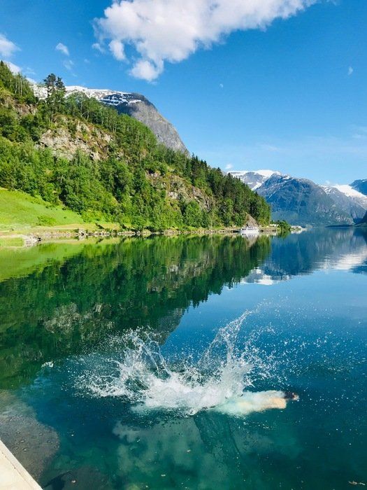 Person som plasker i en turkis alpin innsjø ved siden av grønne fjell og snødekte topper