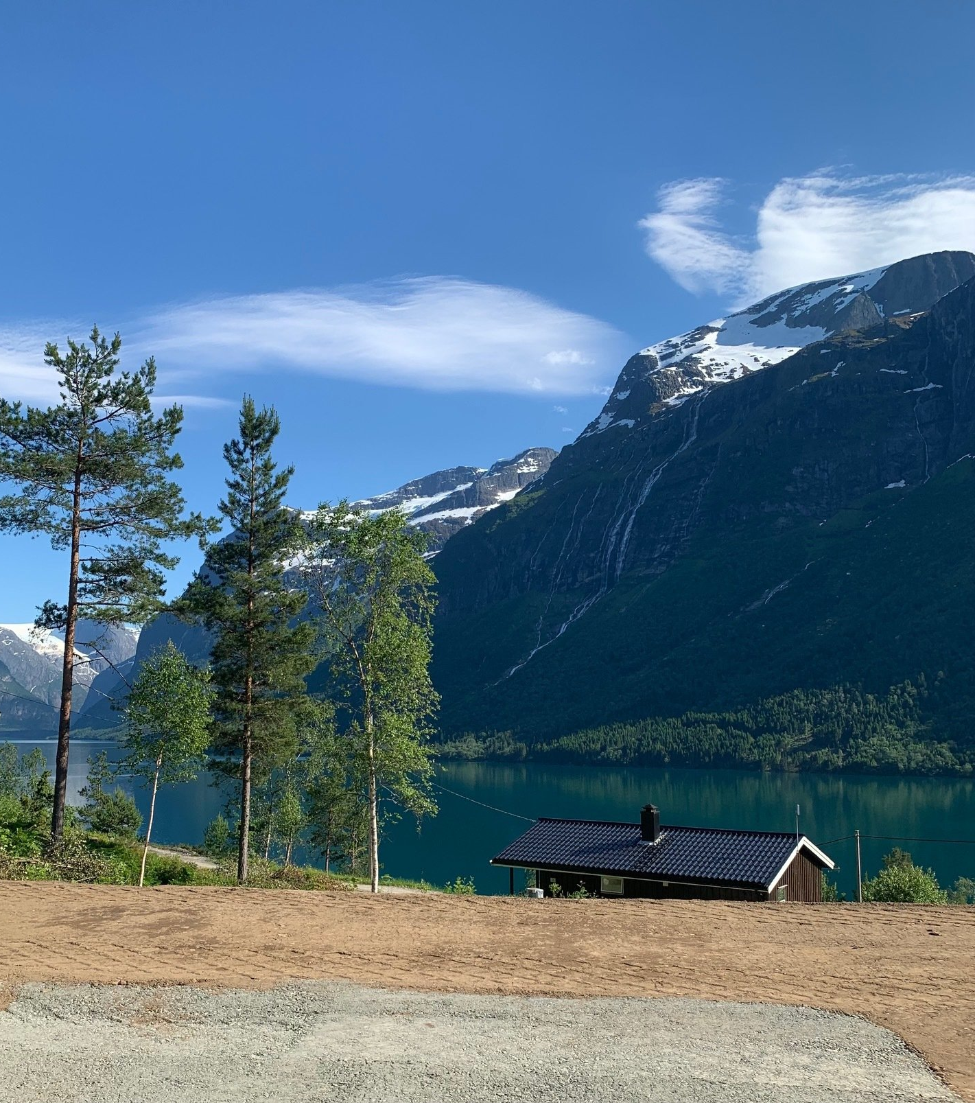 Fjellsjø med snødekte topper, furutrær og en liten hytte under en blå himmel