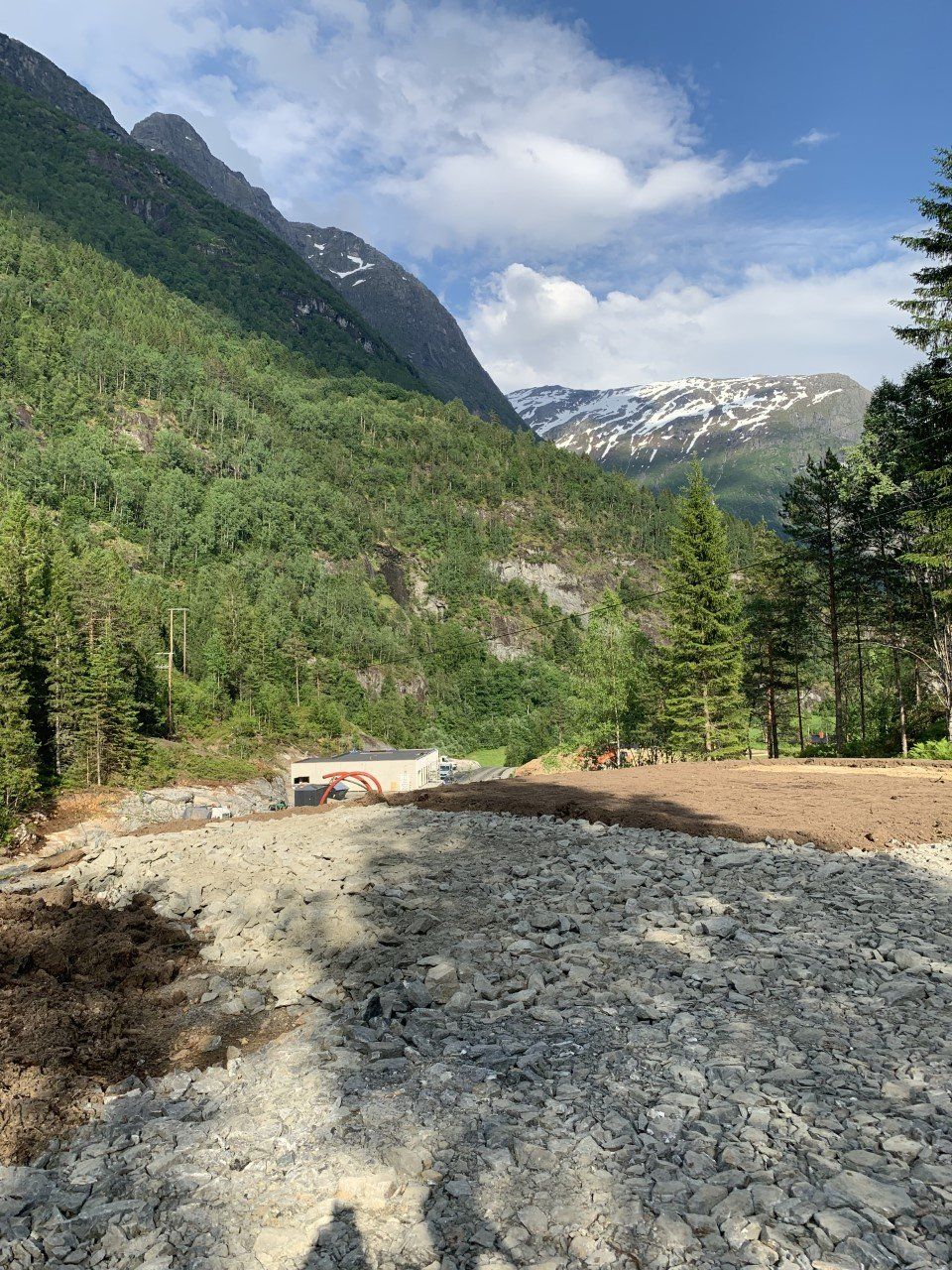 Fjelldal med steinete vei, grønne skogkledde skråninger og snødekte topper under en overskyet himmel