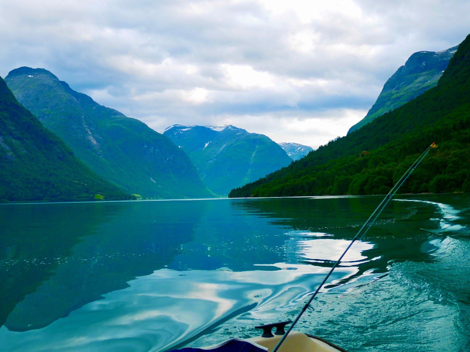 Båtutsikt over en turkis fjord med fjell og overskyet himmel