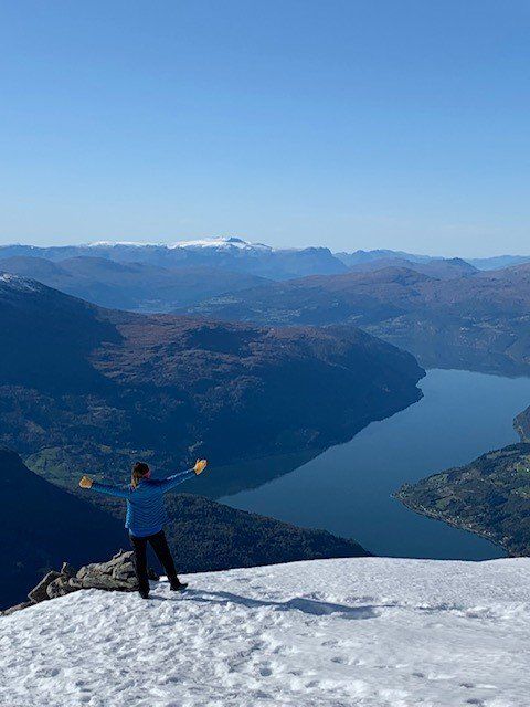 Person som står på et snødekt fjell med utsyn over en innsjø og fjerne topper