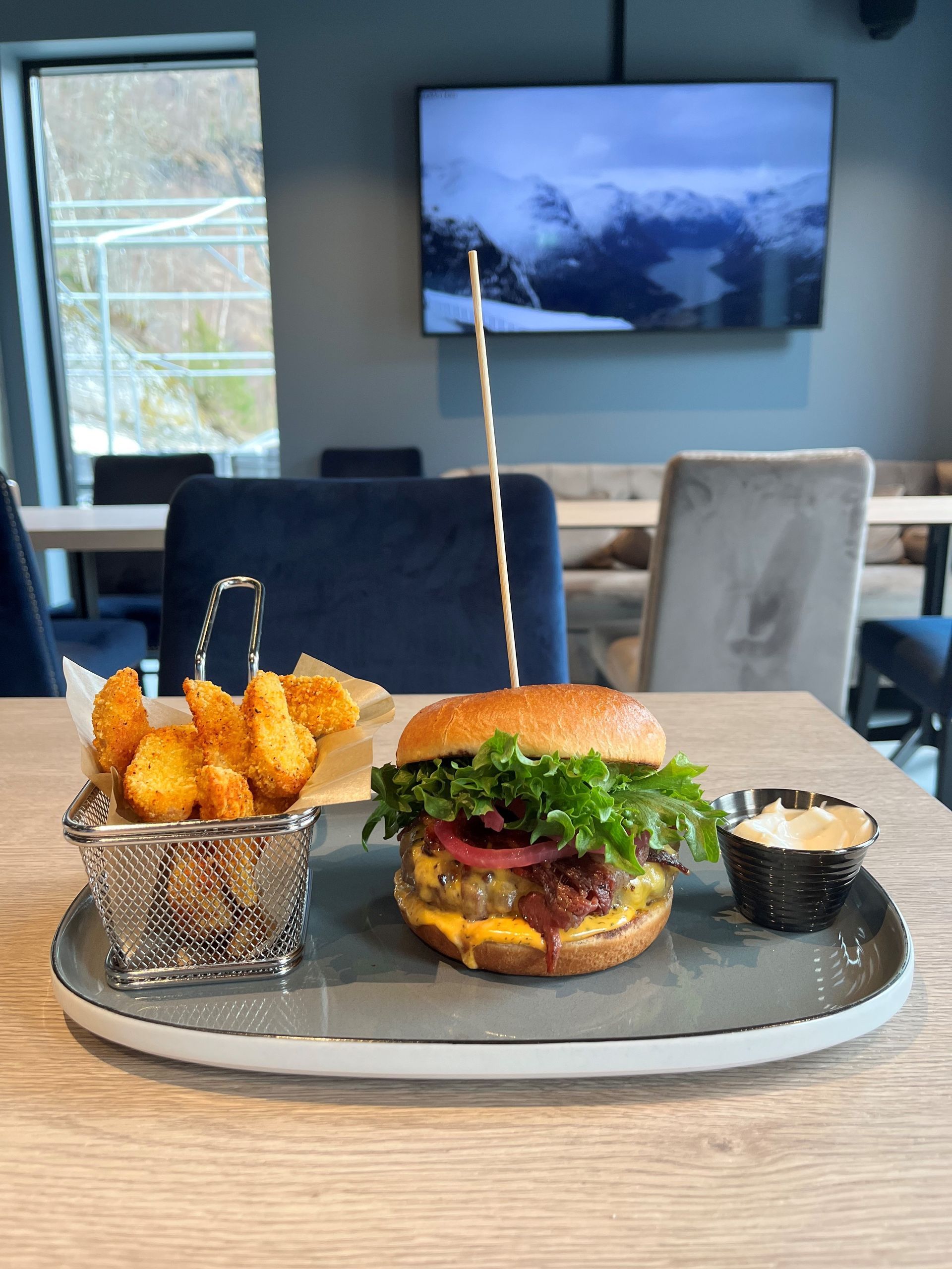Burger, pommes frites og dippsaus på et brett i en moderne kafé med en veggmontert TV i bakgrunnen