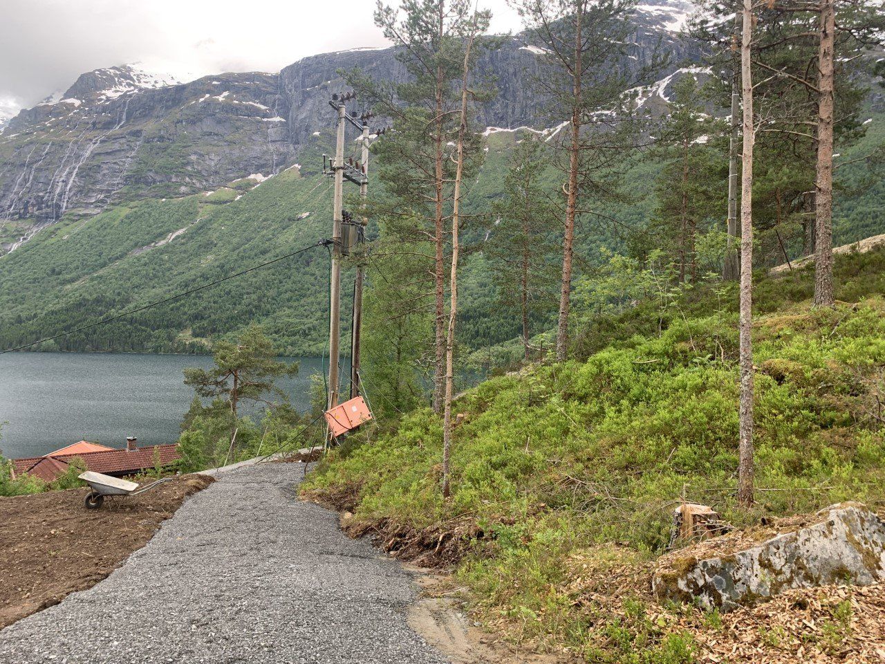 Grussti ved en innsjø med furutrær og bratte grønne fjell under overskyet himmel