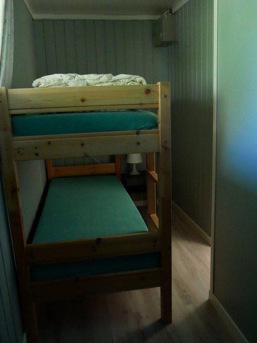Køyeseng med blågrønn madrass i et smalt rom med paneler og et vindu til høyre.