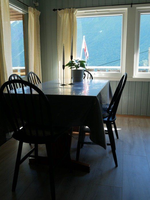 Spisebord og stoler ved lyse vinduer med utsikt over blått vann og en brygge.