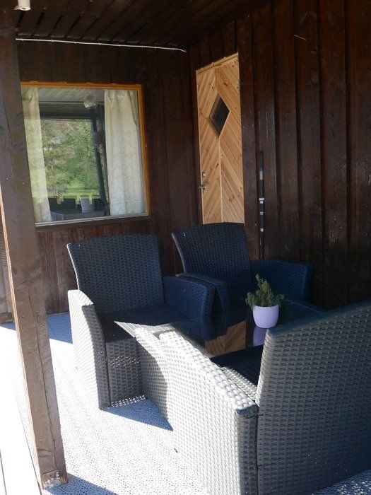 Overbygd veranda med kurvstoler og et lite bord, mørke trevegger og et vindu med gardin