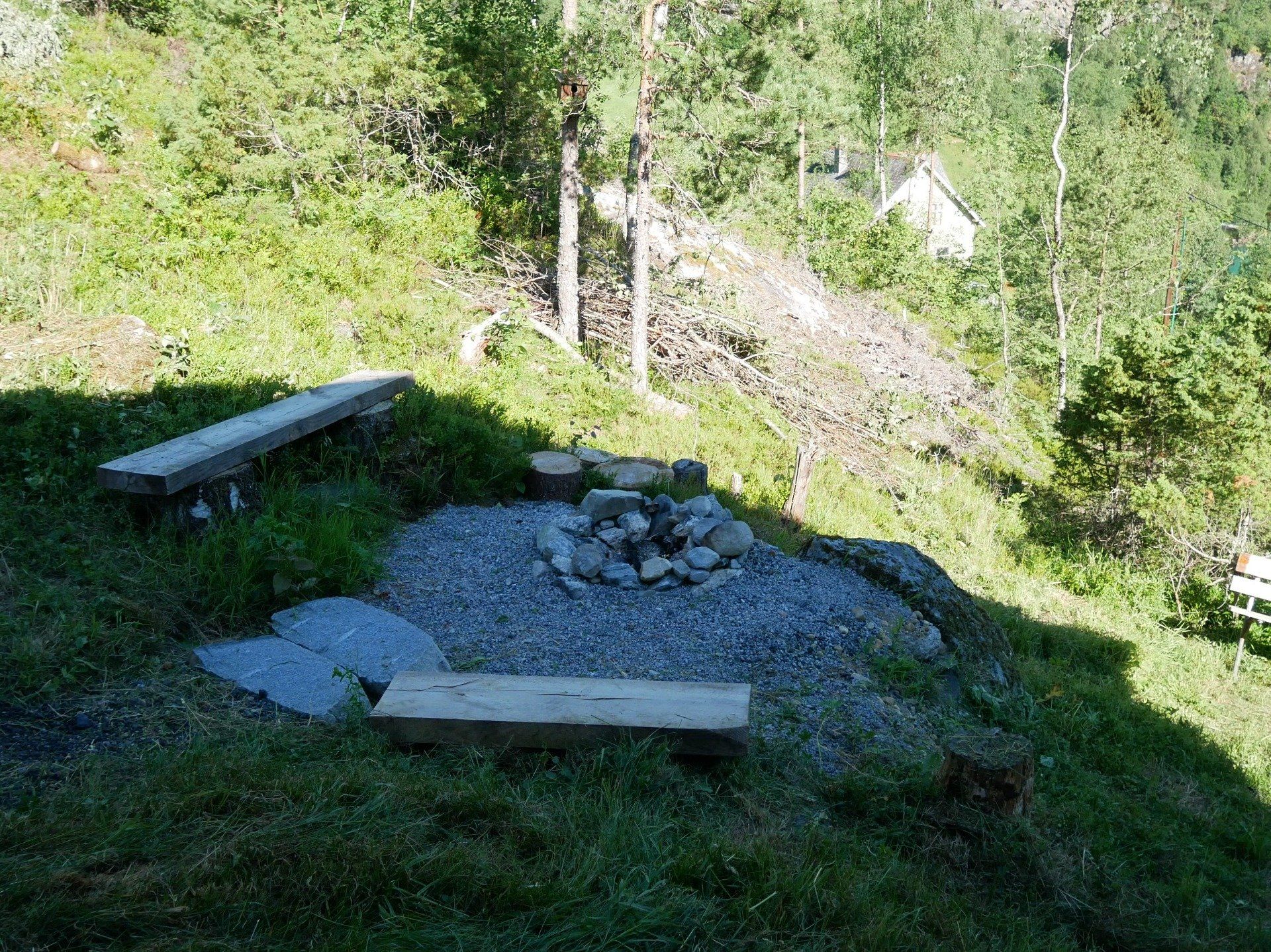 Bålplass i stein med trebenker i en gresskledd åsside, omgitt av trær og steiner.