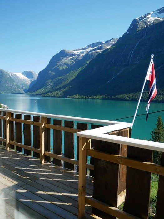 Treterrasse med utsikt over en blå fjord og fjelldal, med et norsk flagg på rekkverket.