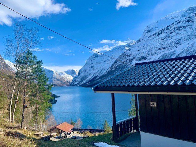 Snødekte fjell ved en blå fjord, med et hyttetak i forgrunnen under en klar himmel