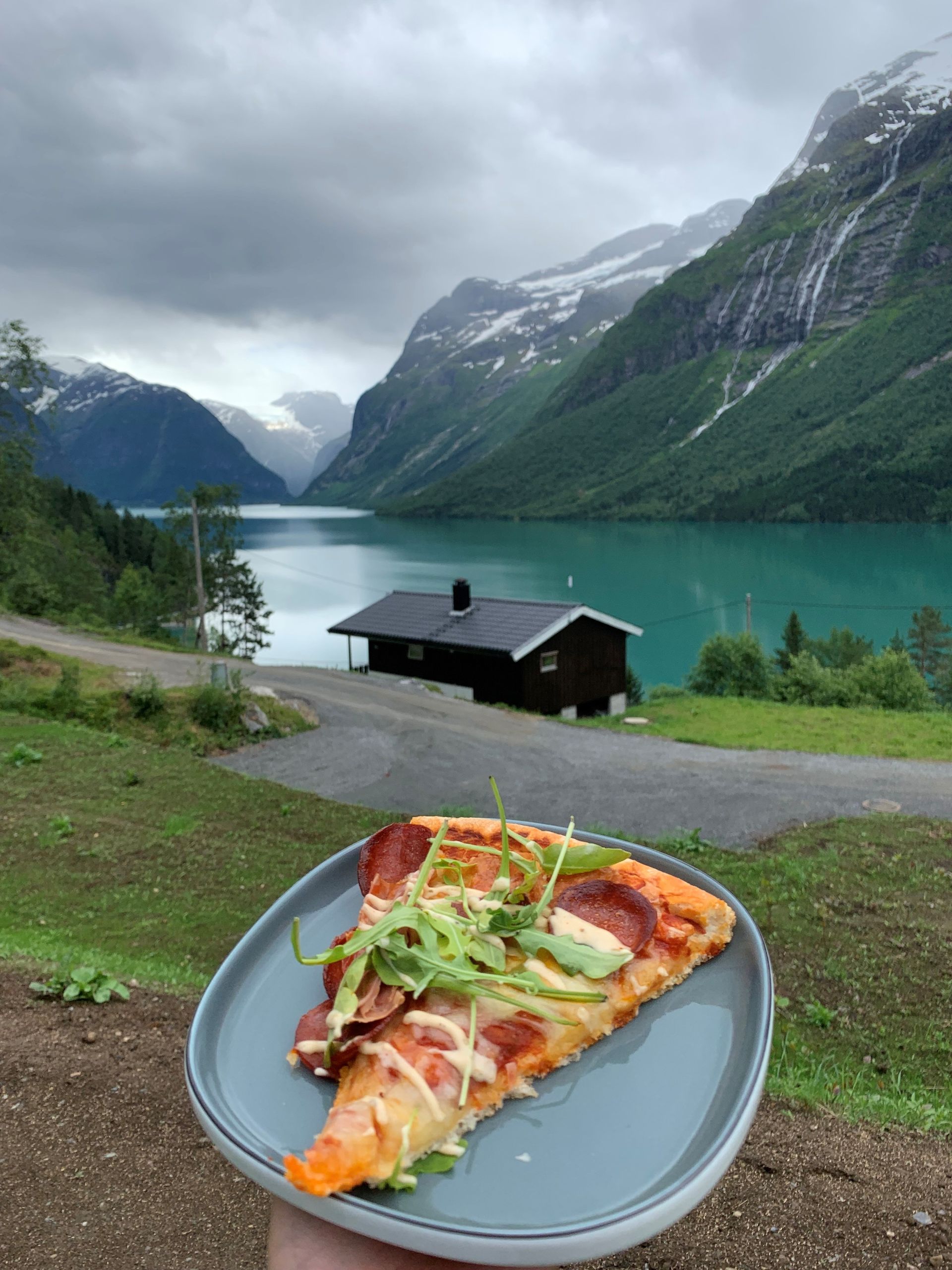 Pizzastykke på en tallerken med utsikt over en turkis fjord, fjell og en liten hytte under overskyet himmel
