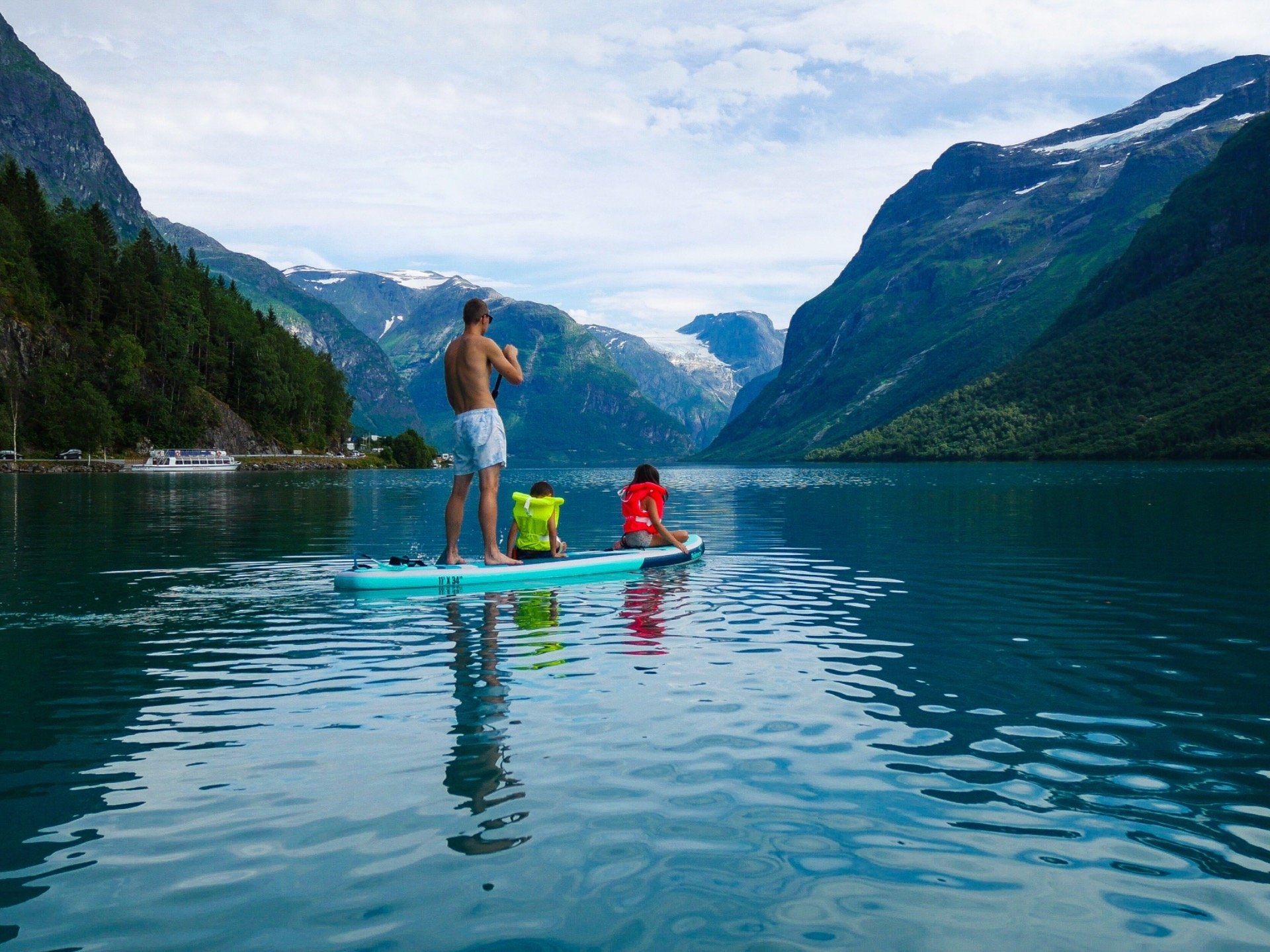 To personer padler på en rolig fjellsjø med skogkledde klipper og snødekte topper i bakgrunnen