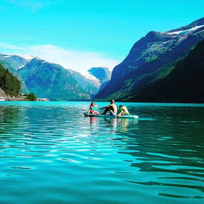Tre kajakkpadlere på turkis vann i en fjellfjord under en lys blå himmel