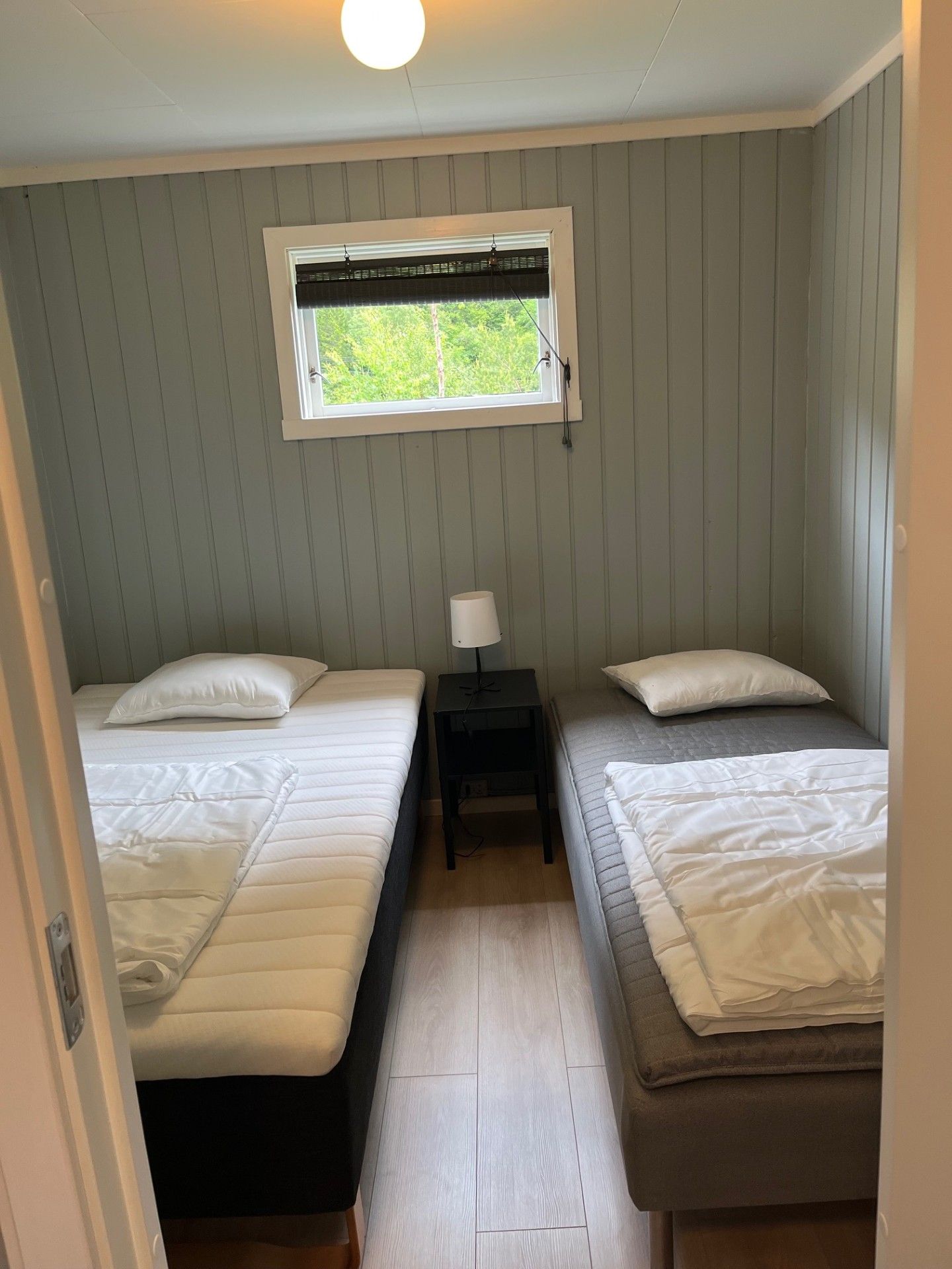 Lite soverom med to enkeltsenger, nattbord og et vindu med grøntområder.