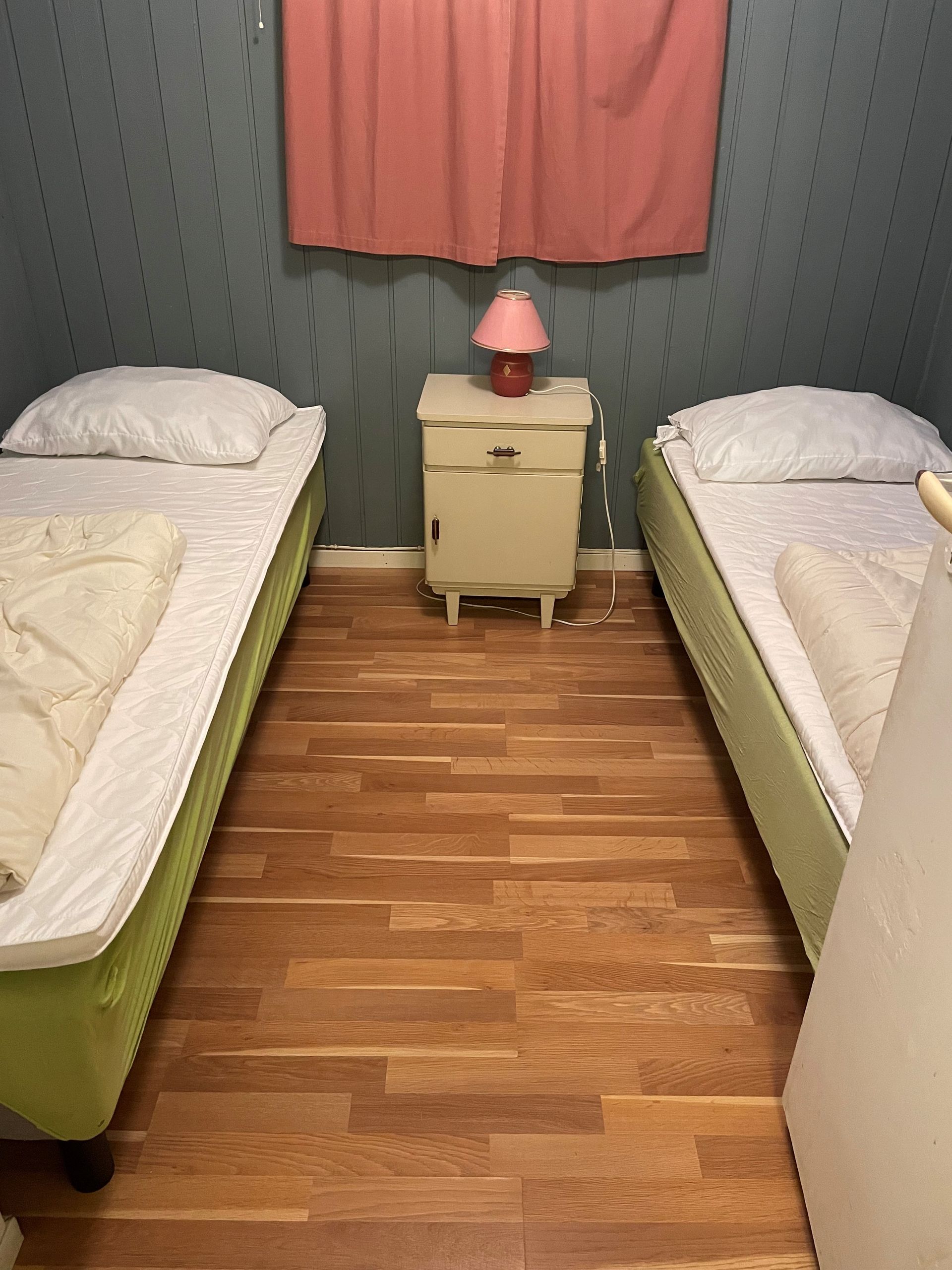 Lite soverom med to enkeltsenger, en nattbordlampe og rosa gardiner mot en mørkegrønn vegg.