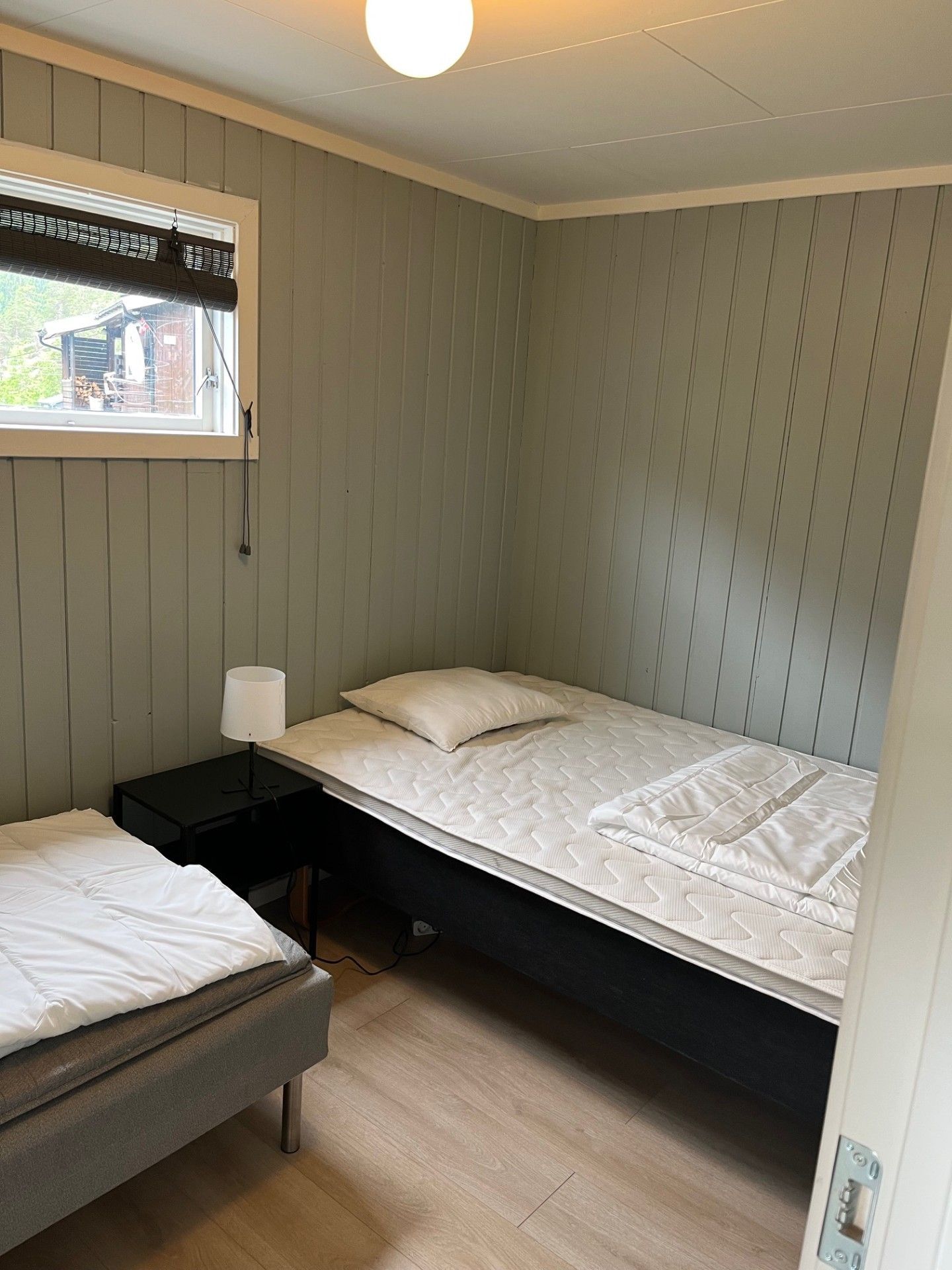 Lite soverom med to enkeltsenger, grå vegger og et vindu som slipper inn dagslys.