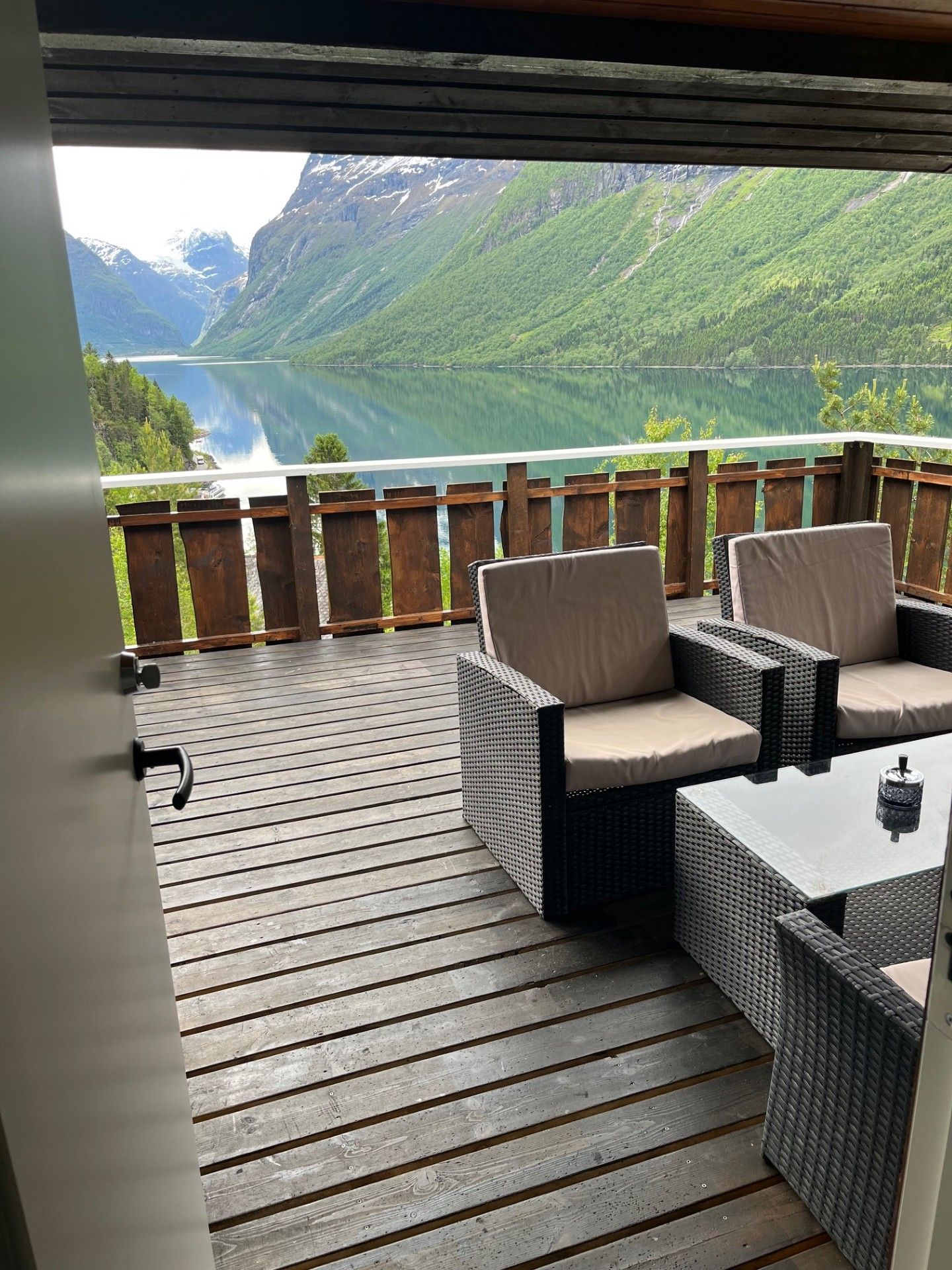 Utsikt over innsjøen og fjellene fra en treterrasse med kurvstoler og et bord under en overbygd terrasse.