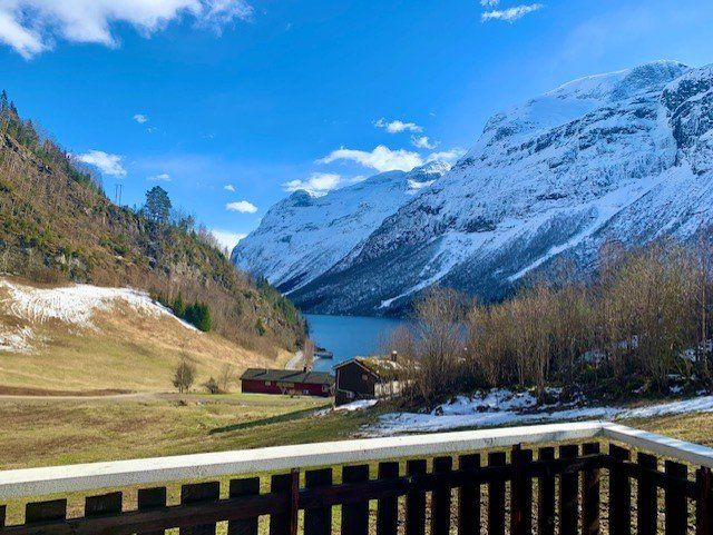 Snødekt fjelldal med en blå fjord, gresskledd mark og rød hytte under en klar himmel