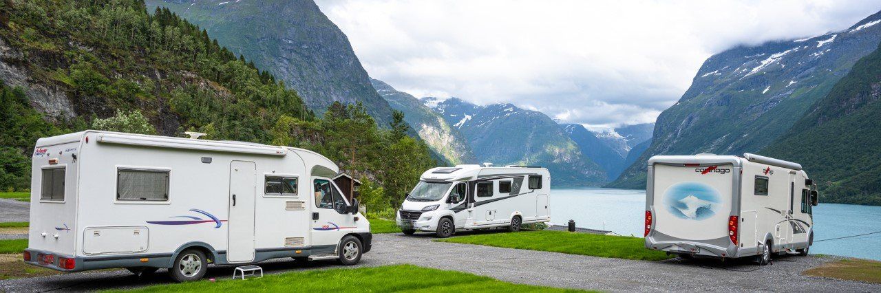 Tre hvite bobiler parkert ved en innsjø med fjell og overskyet himmel i bakgrunnen