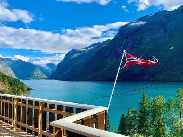 Norsk flagg på et utkikkspunkt over en turkis fjord med fjell og blå himmel