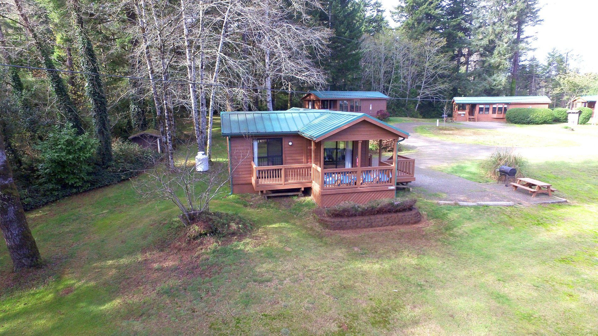 Wooden cabin with blue roof in a grassy yard, surrounded by trees and a picnic area nearby.