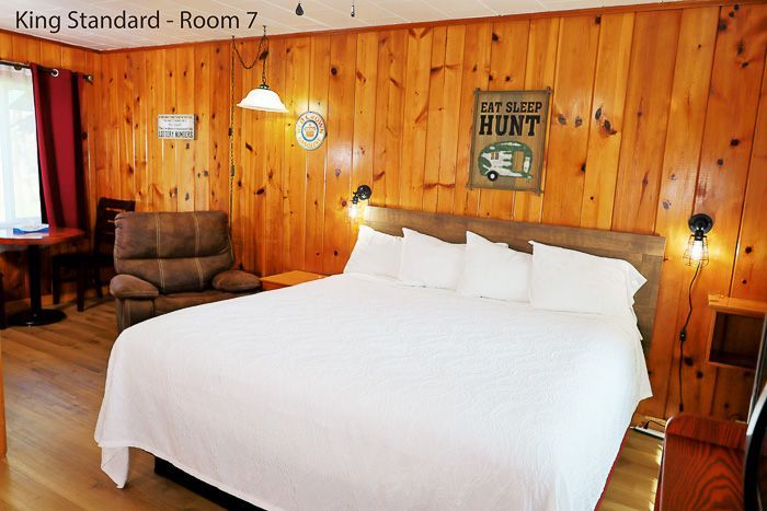 Wood-paneled hotel room with king bed, white linens, and a brown armchair by the window.