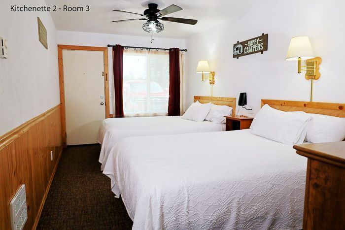 A motel room with two white beds, wood-paneled walls, ceiling fan, and lamps on the nightstands.