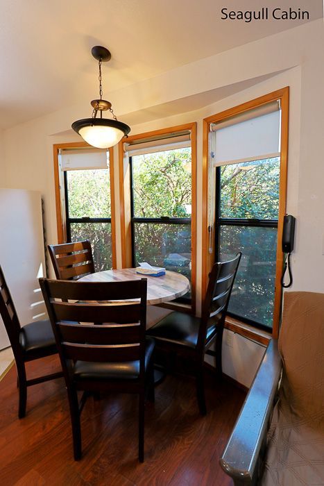Dining nook with round table and wooden chairs beside large corner windows in a sunlit cabin