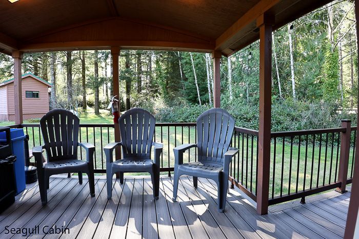 Covered wooden porch with three chairs, black railings, and a wooded yard beyond.