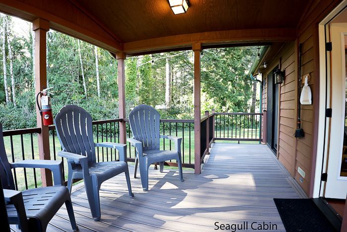 Covered cabin porch with two gray chairs, wooden railing, and trees beyond.