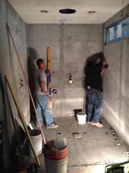 Two workers installing fixtures in an unfinished concrete room with tools and a bucket on the floor