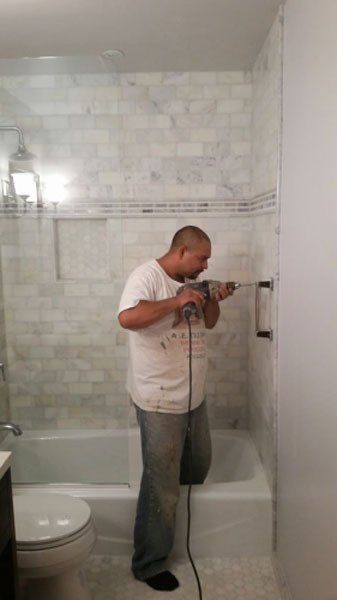 Person drilling a wall in a white-tiled bathroom near a bathtub and toilet