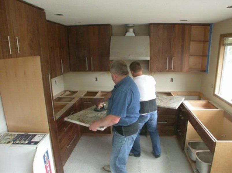 Two men install a countertop in a wood-paneled kitchen under renovation.