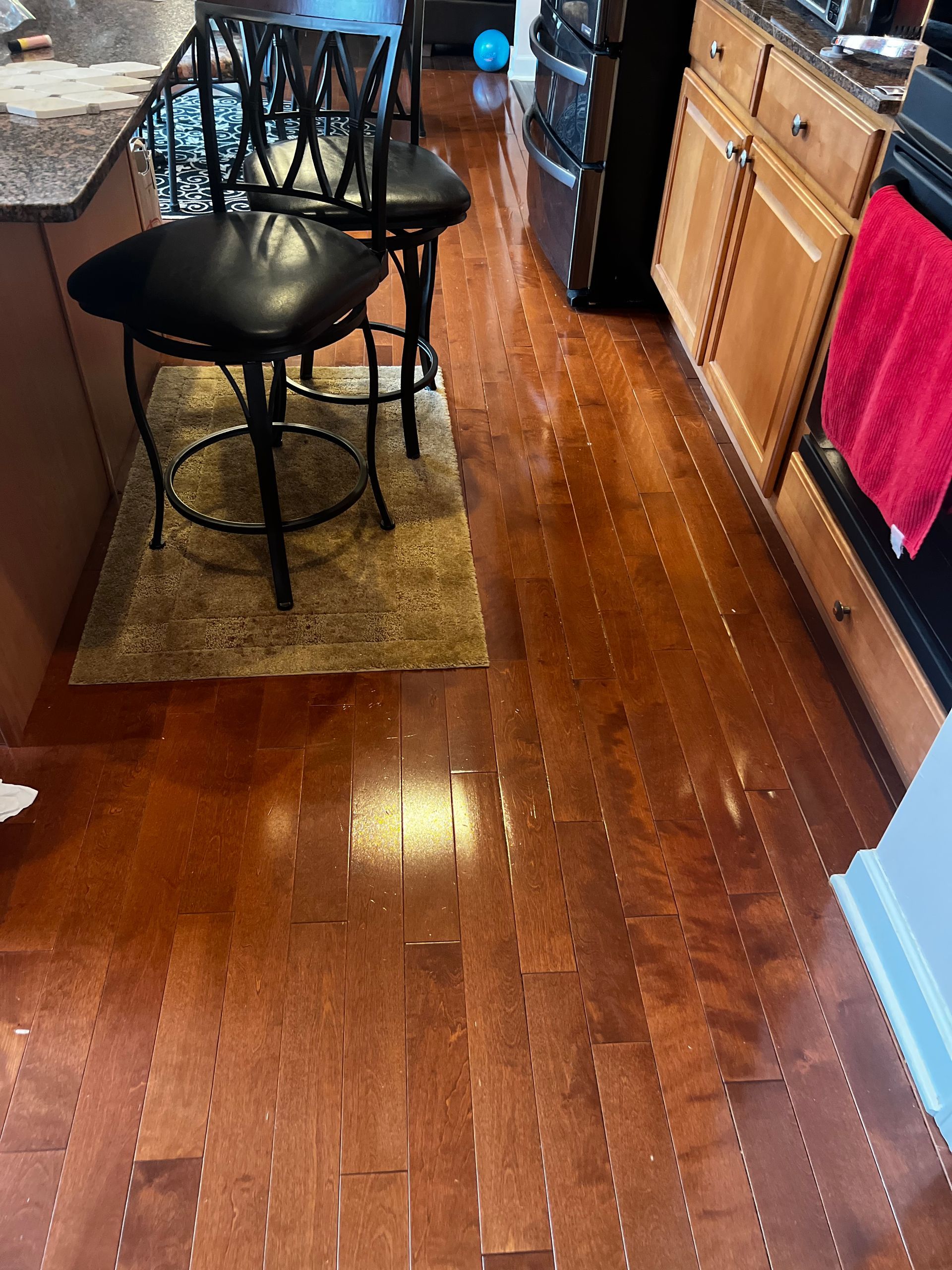 Kitchen with wooden floors, black barstools, and tan cabinets with a pink towel hanging on the oven handle