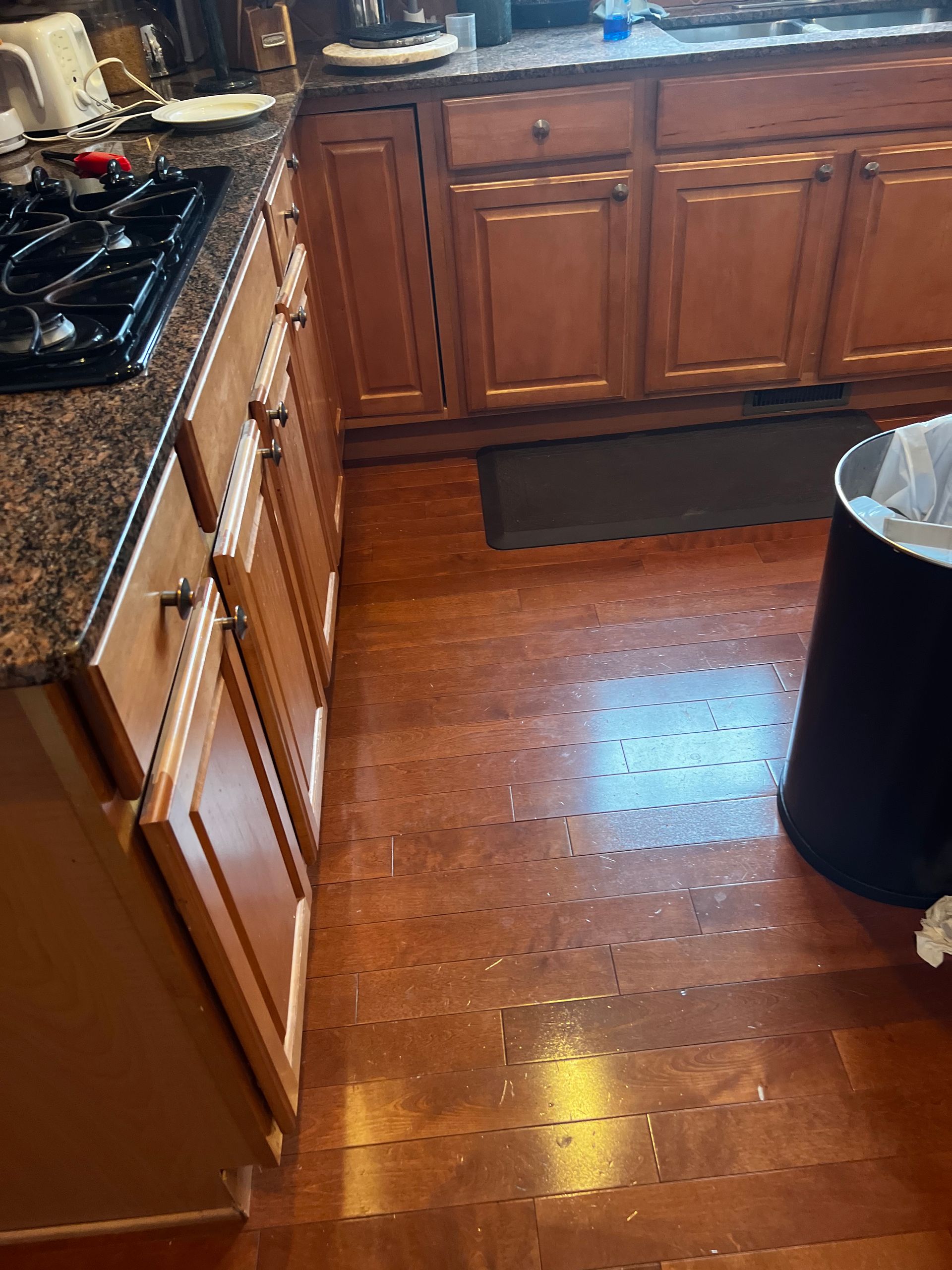 Wooden kitchen floor with cabinets and a stove, plus a black trash can on the right.